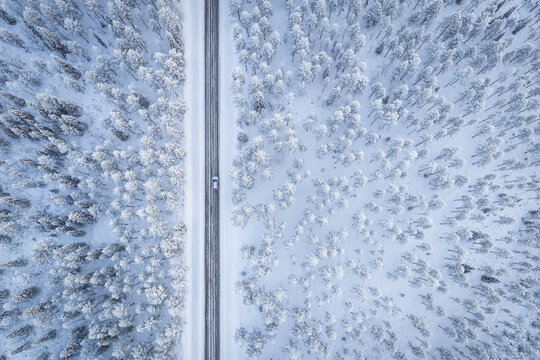 Aerial view of a single car driving on a snow-lined road cutting through a dense, snow-covered forest, Ruka, Pohjois-Pohjanmaa, Finland.