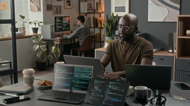 African American young software engineer with beard and glasses typing on laptop at table during workday in modern office of tech company, with two laptops displaying codes in foreground