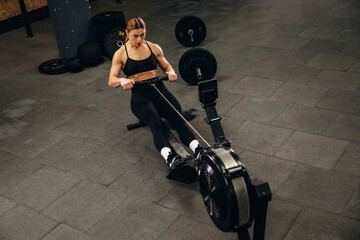 Fototapeta premium Female athlete performing rowing exercise on machine in gym, with weights and fitness equipment visible in the background
