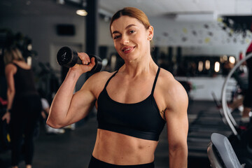 Fototapeta premium Female fitness enthusiast in black sports bra poses with dumbbell in gym, showcasing toned arms and workout equipment in the background
