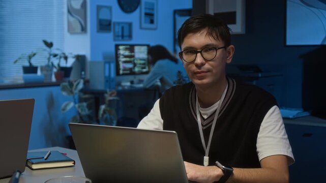 Portrait of Caucasian young male project manager wearing glasses and sweater vest with white t-shirt looking at camera with smile during workday in dark office
