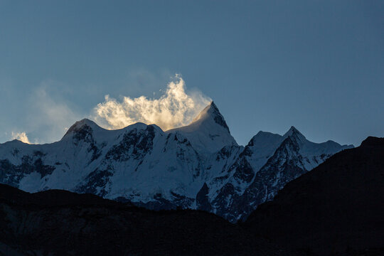 View of jagged, snow-capped peaks pierce the sky, their summits veiled in ethereal clouds touched by the sun's glow, Hunza Nagar, Gilgit, Pakistan.