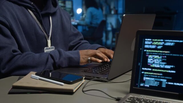 Midsection rack focus shot of unknown Black male web developer wearing blue hoodie and badge programming on laptop at work table with notebooks, pen and smartphone on it