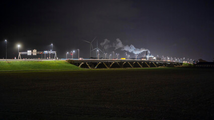 Dutch highway viaduct illuminated at night with wind turbines and industrial steam clouds in the background over green embankment © Victor