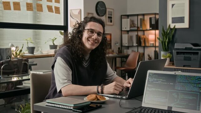 Portrait of positive long-haired gen Z programmer wearing glasses smiling at camera sitting at wooden desk in front of laptop screen in IT office