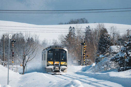 View of a train slicing through a snow-laden path, leaving a trail of powdery snow against the backdrop of stark trees, Biei, Hokkaido, Japan.
