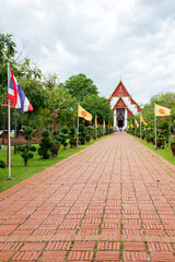 Obraz premium View of the paved ceremonial path leading to the Wihan Phra Mongkhon Bophit in Ayutthaya, Thailand. The path is lined with green bushes and flagpoles flying the Thai national flag and yellow flags.