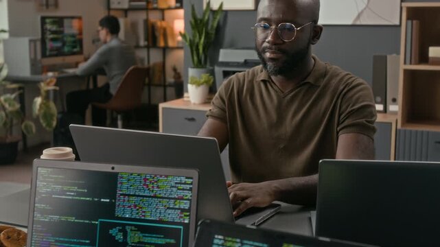 Tilt up view rack focus view of bald Black IT specialist coding and adjusting glasses while working at office table, two laptops with programming codes on screens