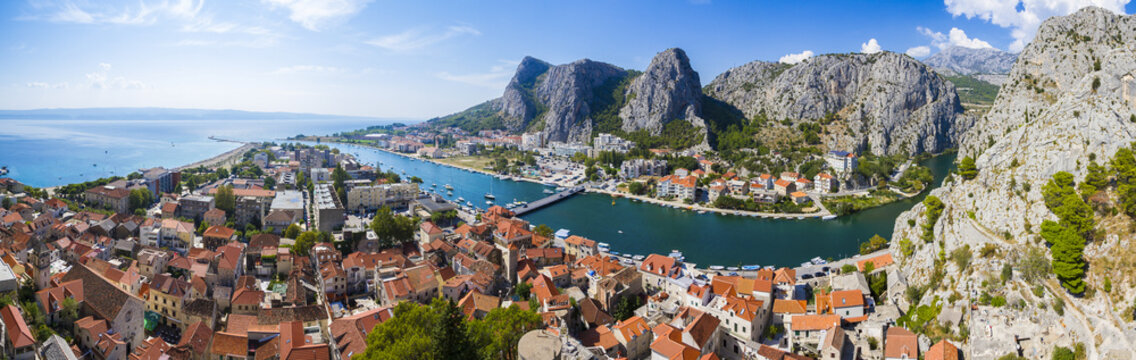 View of terracotta rooftops cascade toward the turquoise river, embraced by towering grey cliffs under a bright blue sky in Omis, Split-Dalmatia County, Croatia.