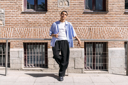 Young man in casual streetwear leaning on a railing by a brick building, exuding modern urban style, confidence and youth culture