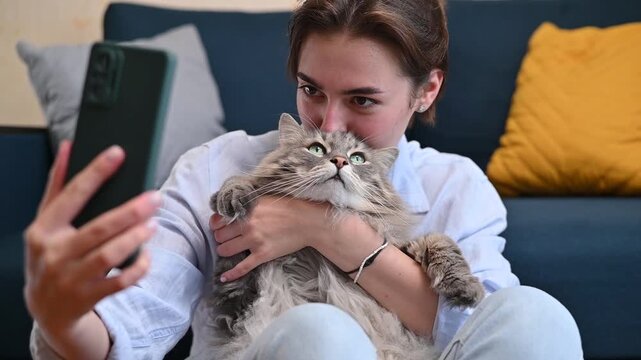 A woman sits on a sofa holding a cat while taking selfies with a smartphone. She smiles and engages with the camera.