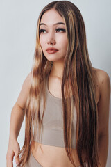 Naklejka premium Young woman with long straight hair and neutral makeup wearing beige crop top standing against light background with serious expression looking aside.