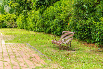 Fototapeta premium Wooden bench with ornate metal frame placed on lawn beside brick path, surrounded by dense green hedges in public park setting.