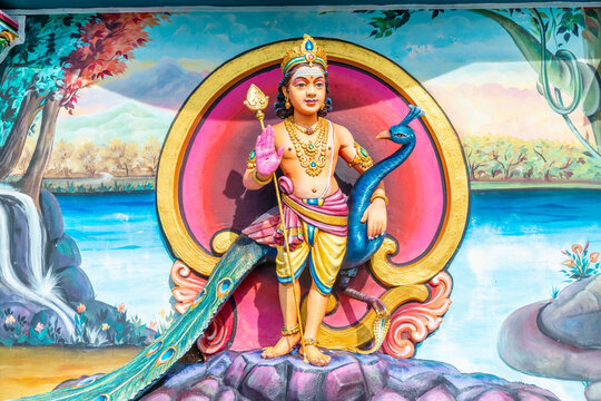 Lord Murugan statue holding spear and blessing hand gesture standing with peacock vahana, colorful painted mural background at Hindu temple, religious art and cultural heritage.