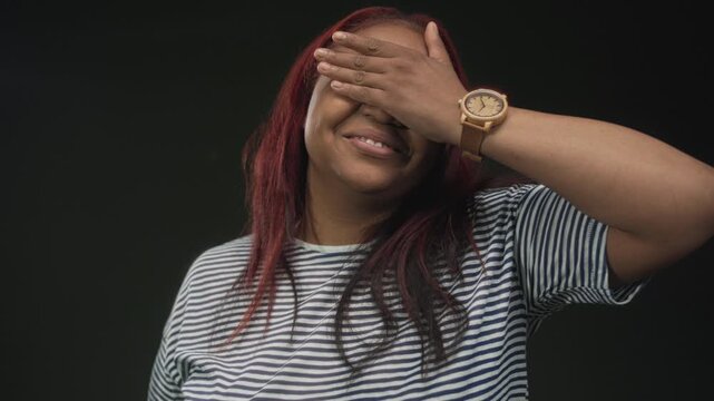 Woman wearing striped shirt covers eyes with hand in dark green studio setting; secrecy concealment embarrassment.