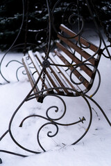 A wooden garden swing with decorative wrought iron swirls sits covered in a fresh layer of white snow in a quiet park during winter, evoking a peaceful and cold seasonal atmosphere.