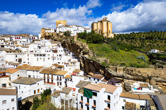 View of clustered white buildings nestled into rocky cliffs, topped by historic structures against a vibrant blue sky, Setenil de las Bodegas, Andalusia, Spain.