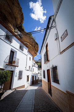 View of whitewashed buildings nestled under a massive overhanging rock formation, contrasting sharply against the bright blue sky, Setenil de las Bodegas, Andalusia, Spain.