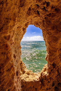 View of turquoise ocean waters shimmer through a rough-hewn cave opening carved into golden sandstone cliffs, a glimpse of paradise, Gruta da Boneca, Portugal.
