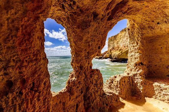 View of sunlight kisses the rugged, golden cliffs framing the azure sea through a natural rock window, Gruta da Boneca, Faro, Portugal.