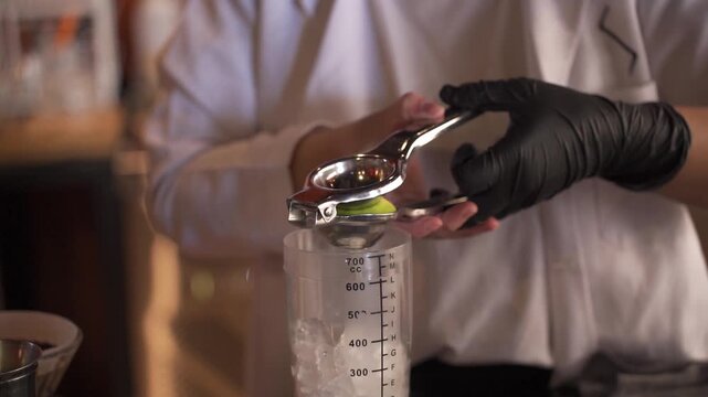 A professional bartender in a white uniform and black gloves uses a metal citrus press to squeeze fresh lime juice into a graduated plastic shaker filled with ice cubes while preparing a cocktail.