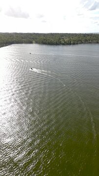 Aerial view of a jet ski rapidly crossing the sunlit surface of Lake Dyer, leaving a prominent wake behind while surrounded by lush green Australian landscape and a partly cloudy sky