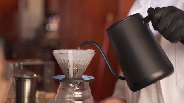 A close-up of a professional barista's hands in black gloves carefully pouring hot water from a gooseneck kettle into a V60 coffee dripper to brew artisanal specialty coffee.