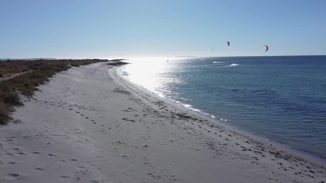 Beautiful aerial drone shot flying over a sandy dune path and revealing a sunlit turquoise ocean with several kitesurfers enjoying a perfect windy day on a stunning coastline