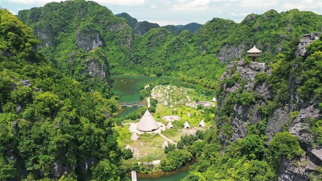 A stunning high-angle aerial shot reveals the Kong movie set with thatched huts and a large central conical structure, hidden among the spectacular limestone mountains of Trang An.