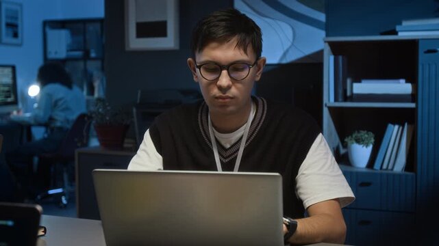 Concentrated Caucasian gen Z male quality assurance specialist adjusting glasses and typing on laptop while sitting at office desk at nighttime