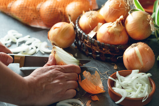 Corte de cebolla y escena activa de preparaci&oacute;n en cocina.Manos cortando una cebolla amarilla sobre encimera oscura, rodeada de cebollas enteras, cesta y cuenco. Momento culinario en plena preparaci&oacute;n