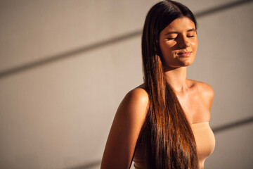 Young woman with long brown hair stands in soft light, eyes closed, enjoying tranquility, with gentle shadows creating a serene atmosphere and highlighting her natural beauty