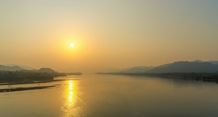 Obraz premium Yellow sun rising on serene river with distant blue mountains beneath clear sky