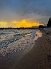 Fototapeta premium Empty beach at Issyk Kul Lake in Kyrgyzstan before a storm. Dark clouds and rain over the water with golden sunset glow.
