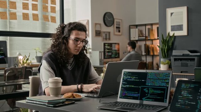 Long-haired gen Z male software engineer wearing glasses working on laptop at grey office table with notebooks, phone, coffee cup, cookies on plate and other laptops on it