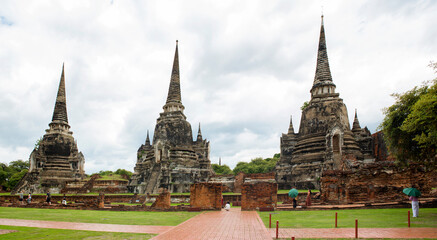 Naklejka premium The three iconic restored chedis (stupas) at Wat Phra Si Sanphet in Ayutthaya, Thailand. The massive, bell-shaped structures stand in a row on a raised platform covered in green grass. Cloudy day.
