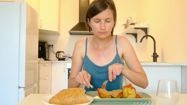 Woman sits at kitchen table eating stuffed paprika, enjoying homemade Balkan cuisine, turning meal into cultural moment shared in quiet domestic space.