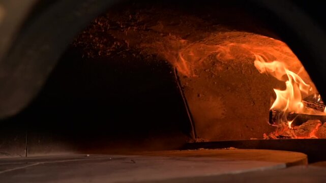 Close-up video of a rotating wood fired pizza oven with active flame inside. The oven floor slowly spins while the fire burns brightly, creating an authentic atmosphere of traditional Italian pizzeria
