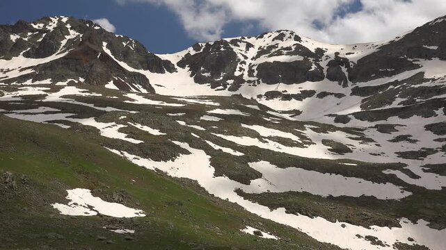 Spring alpine meadow with melting snow on mountain slopes, USA landscape beneath clouds. Vernal highland grassland amid patchy drifts across Rockies ridges under cumulus sky panorama.
