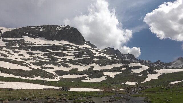 Timelapse clouds drift above snow patches on Rocky Mountains, spring alpine meadow, USA. Fast moving sky rolls past rugged ridges and green grassland as melting drifts fade in America.