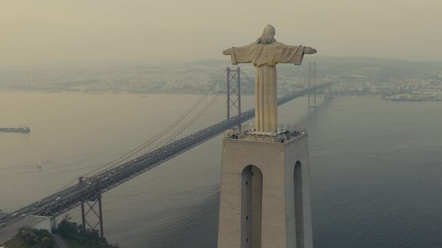 Drone view of the Christ the King monument in Almada, Portugal, with the 25 de Abril Bridge spanning the Tagus River and Lisbon visible in the background.
