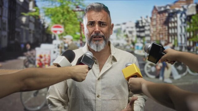 Man with gray beard speaking into multiple microphones on a city street while gesturing with his hands; concern accountability.