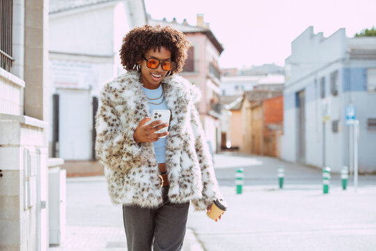 Latin american black woman with curly hair walking urban street, smiling using phone and holding coffee.