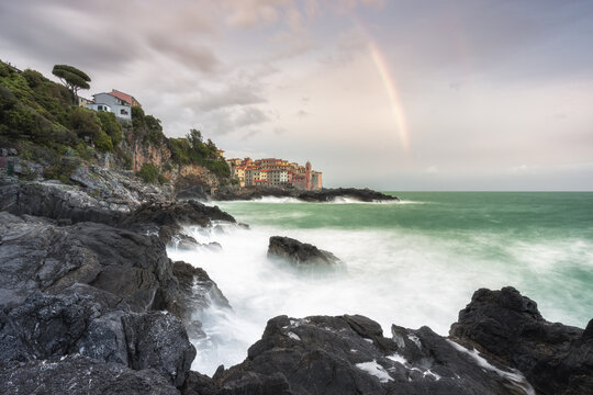 View of waves crashing against the rocky shore under a pastel sky with a faint rainbow arcing over the village, Tellaro, Liguria, Italy.
