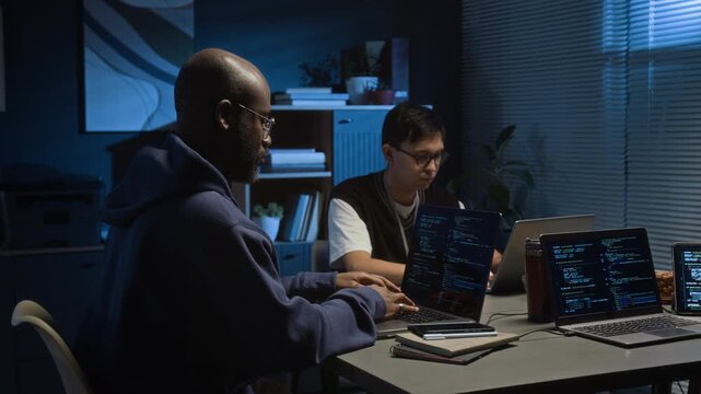 Two diverse young male software testers working on laptops in office of IT company at night, Black man removing glasses to rub his tired eyes