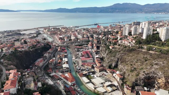 Drone aerial flying above the Rječina River through Rijeka toward the Kvarner Bay and the Adriatic Sea on a sunny day. Cityscape spreading below with coastline and bay in the distance.