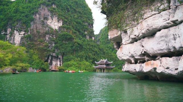 Visitors on traditional rowboats navigate the emerald waters of Trang An, passing by a historic water pavilion and towering limestone cliffs in Ninh Binh, Vietnam.