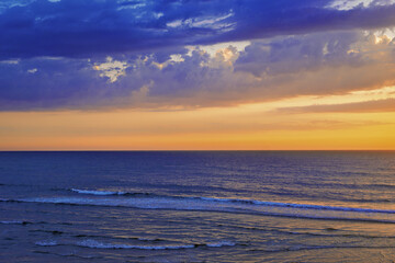 A vibrant sunset spreads above Leirosa Beach, the sky shifting from deep blue and purple to bright orange and gold as clouds catch the last light.