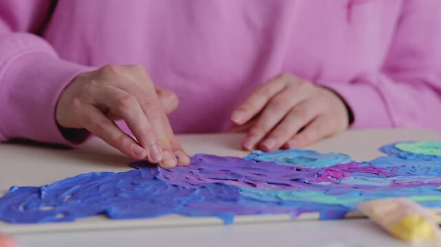 A person uses their fingers to spread blue and purple paint on a canvas. The setting is indoors with good lighting in the afternoon