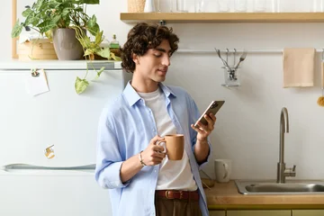 Handsome young man with curly hair enjoying coffee while checking his phone at home © LIGHTFIELD STUDIOS